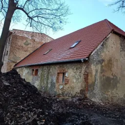 Odnowiony dach z czerwonej dachówki na starym, otynkowanym budynku z widocznymi ceglanymi elementami, z kupą ziemi u podstawy i drzewem z gołymi gałęziami w tle.