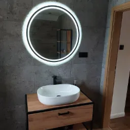 Nowoczesna łazienka z umywalką nablatową na drewnianej szafce z czarnymi elementami, okrągłe lustro z podświetleniem LED na tle ściany wyłożonej szarymi płytkami imitującymi kamień, widoczny...