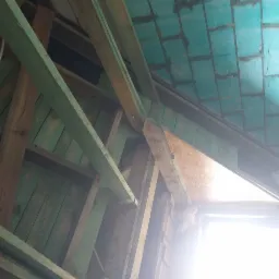 Ocieplenie poddasza z widoczną konstrukcją drewnianą i izolacją w postaci płyt z zieloną warstwą wierzchnią, widok od dołu w kierunku okna.