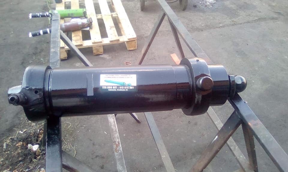 Czarny, odnowiony cylinder hydrauliczny na metalowym stojaku, z naklejoną etykietą firmy, widoczne elementy mocujące i ślady obróbki.