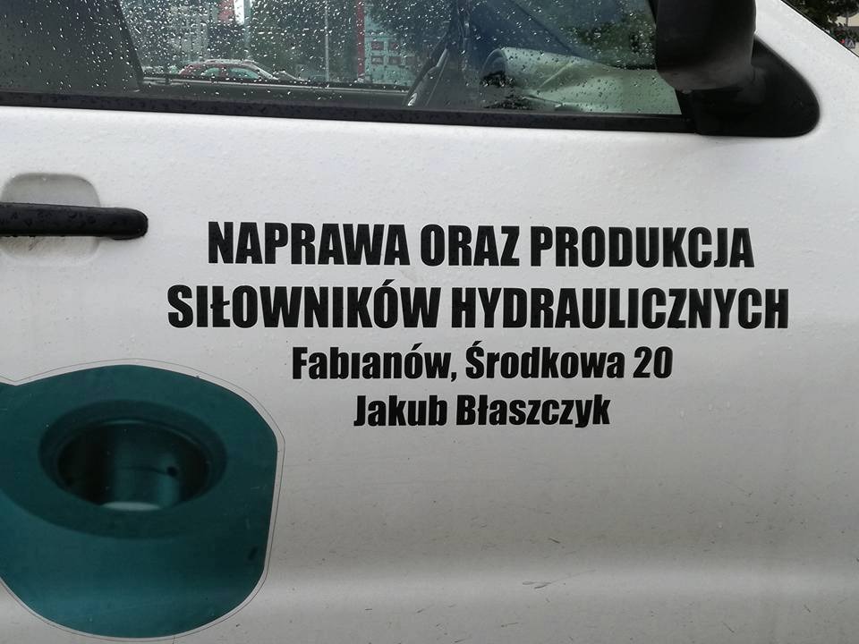 Napis na samochodzie dostawczym informujący o naprawie i produkcji siłowników hydraulicznych, wraz z adresem firmy i imieniem właściciela, widoczne krople deszczu na karoserii.