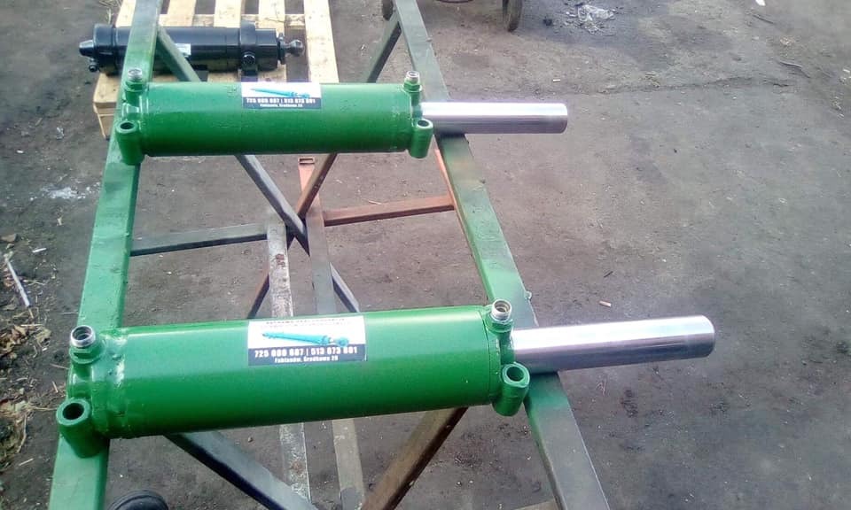 Dwa zielone cylindry hydrauliczne, z chromowanymi tłokami, umieszczone na metalowej konstrukcji pomalowanej na zielono, w tle paleta i inny cylinder.