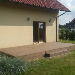 Nowoczesny, brązowy taras kompozytowy z jednym stopniem, przylegający do beżowego domu z brązowymi oknami, otoczony trawnikiem i ozdobną trawą, widok na sąsiedni dom w tle.