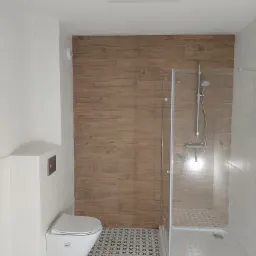 Nowoczesna łazienka z podwieszaną toaletą, kabiną prysznicową ze szklanymi drzwiami, płytkami imitującymi drewno na ścianie i wzorzystą posadzką z motywem krzyżyków.