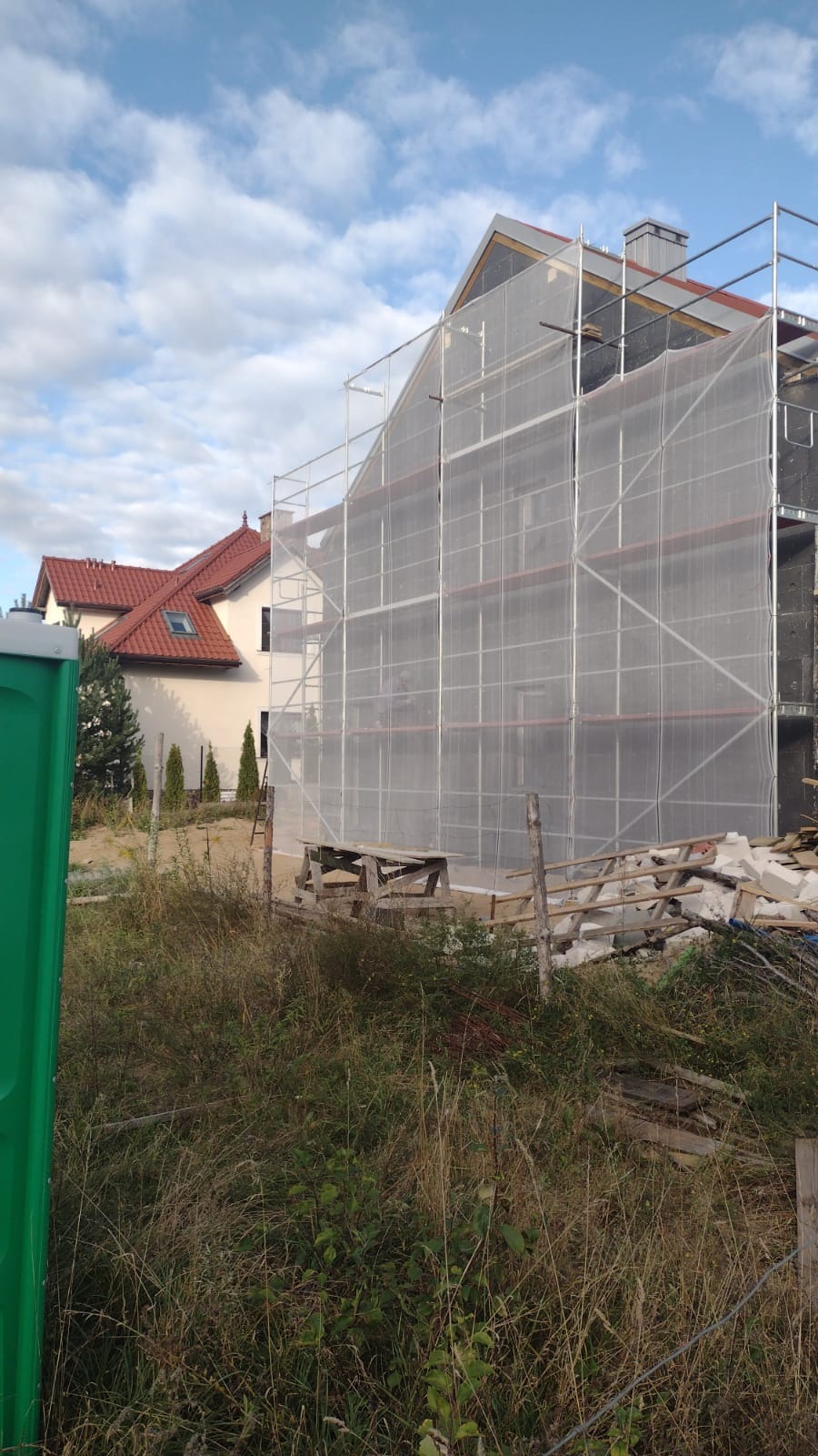 Dom w trakcie ocieplania elewacji, osłonięty siatką na rusztowaniu, widoczny fragment sąsiedniego domu z czerwoną dachówką i zielona toaleta przenośna na pierwszym planie.