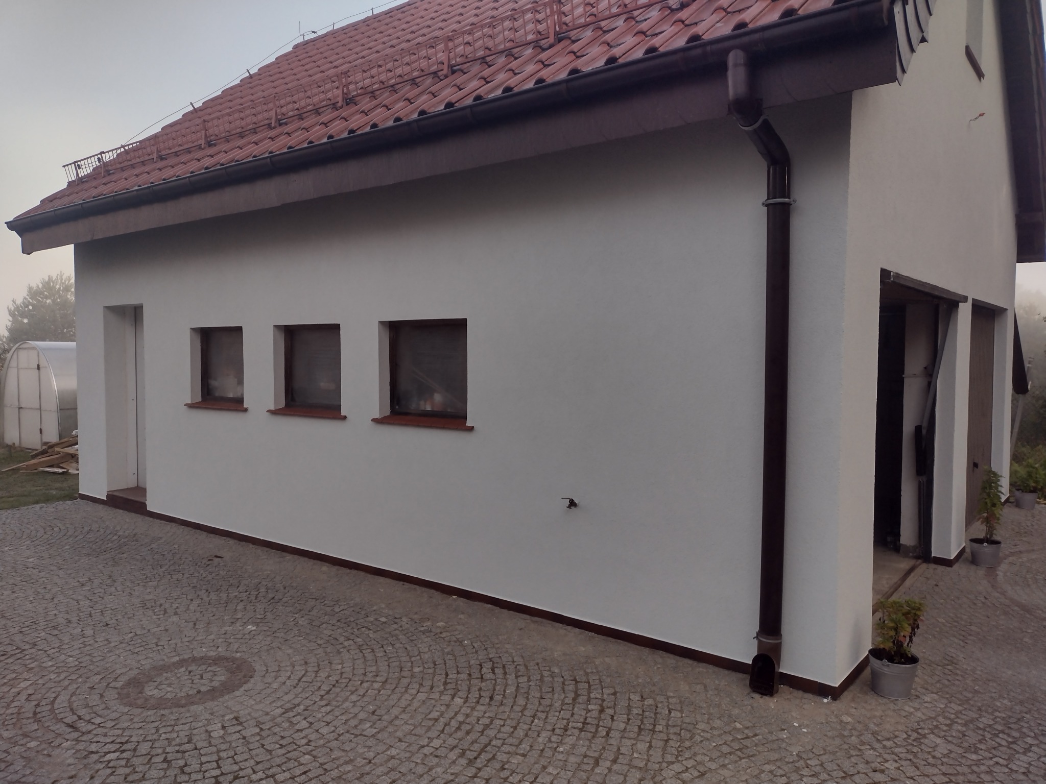 Otynkowany budynek z brązowymi rynnami i czerwoną dachówką, widok z zewnątrz, kostka brukowa na podwórzu, krzewy w donicach przy wejściu do garażu.