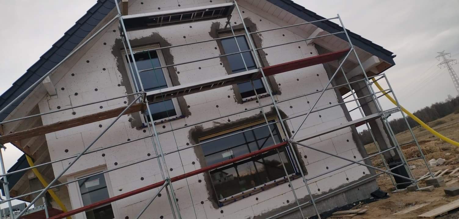 Dom jednorodzinny w trakcie ocieplania styropianem, widoczne rusztowanie i obramowania okien przygotowane do dalszych prac wykończeniowych.