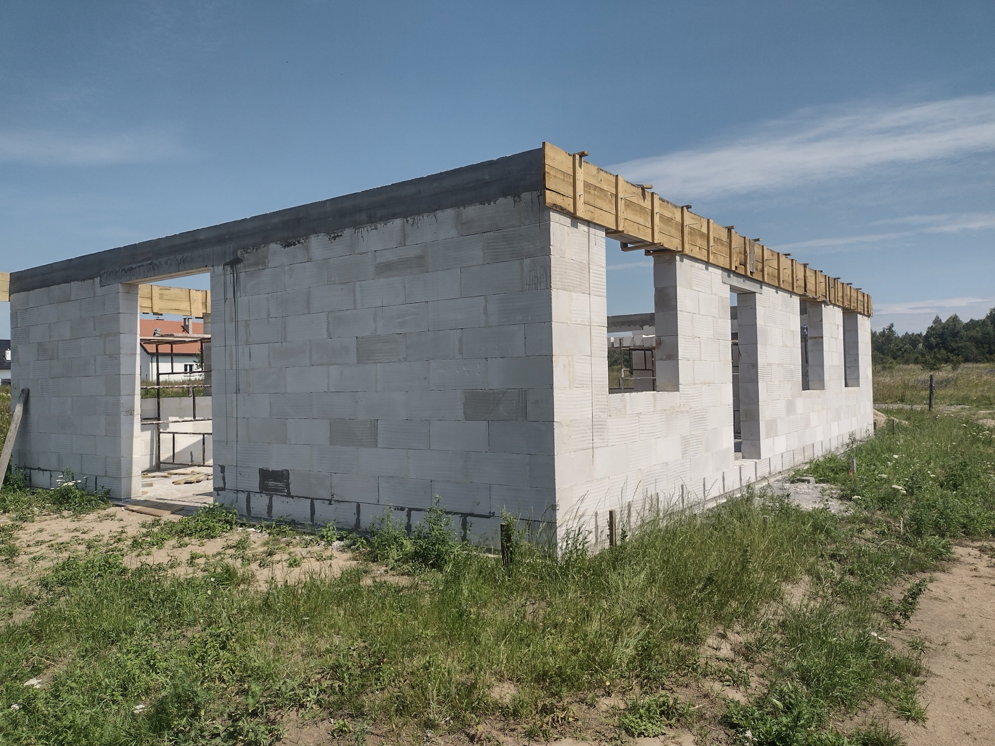 Niedokończony budynek z białych bloczków, widoczne drewniane szalunki na górze, otoczenie zarośnięte trawą, niebo bezchmurne.