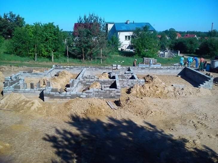 Wykopane fundamenty domu z częściowo wzniesionymi ścianami z bloczków betonowych. W tle widoczny budynek mieszkalny i drzewa. Na budowie obecni pracownicy.
