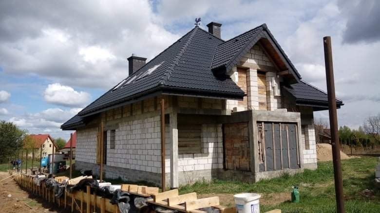 Budowa domu jednorodzinnego z białych bloczków, z czarną dachówką i lukarnami, na etapie murowania ścian i montażu więźby dachowej, widoczny szalunek fundamentów.