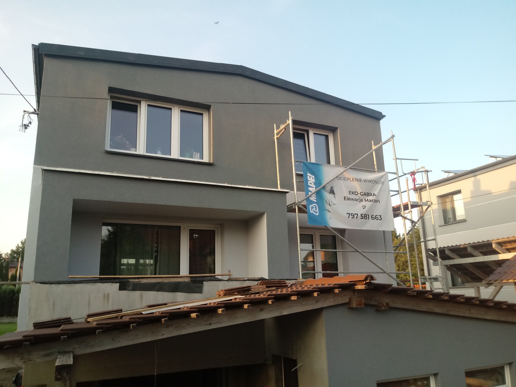 Szary, dwupiętrowy dom w Olsztynie z rusztowaniem i banerem reklamowym firmy oferującej ocieplenie i elewacje, widok na fragment dachu z czerwonej dachówki.