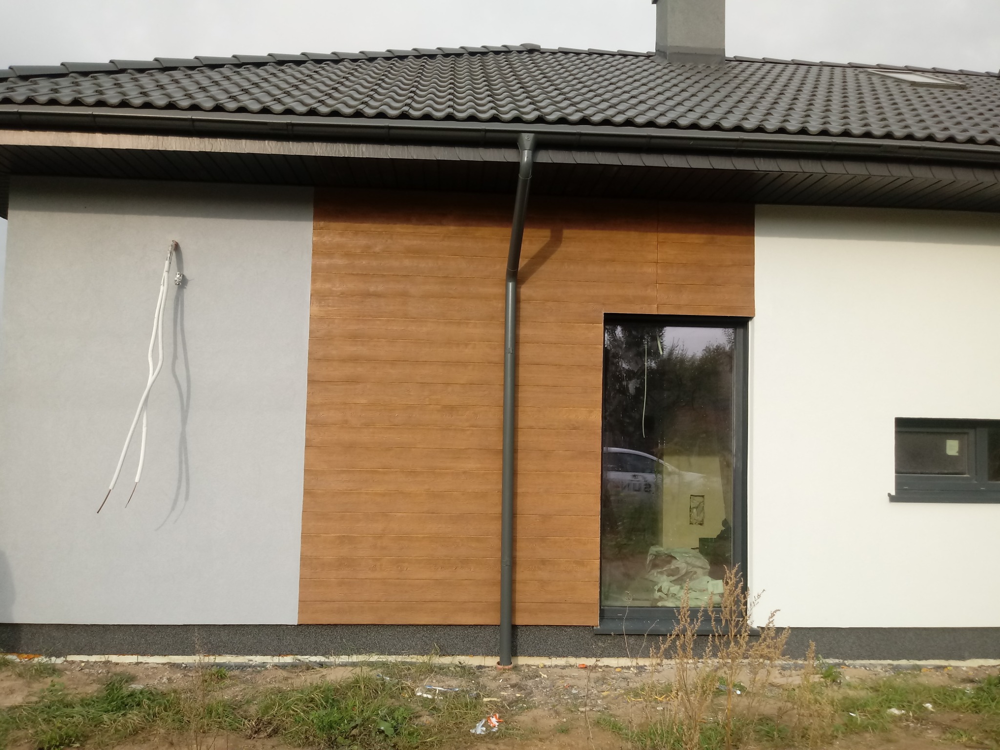 Fragment elewacji domu z trzema rodzajami wykończenia: szary tynk, drewniana okładzina i biała farba, widoczne wystające kable elektryczne, rynna i okno z odbiciem samochodu.