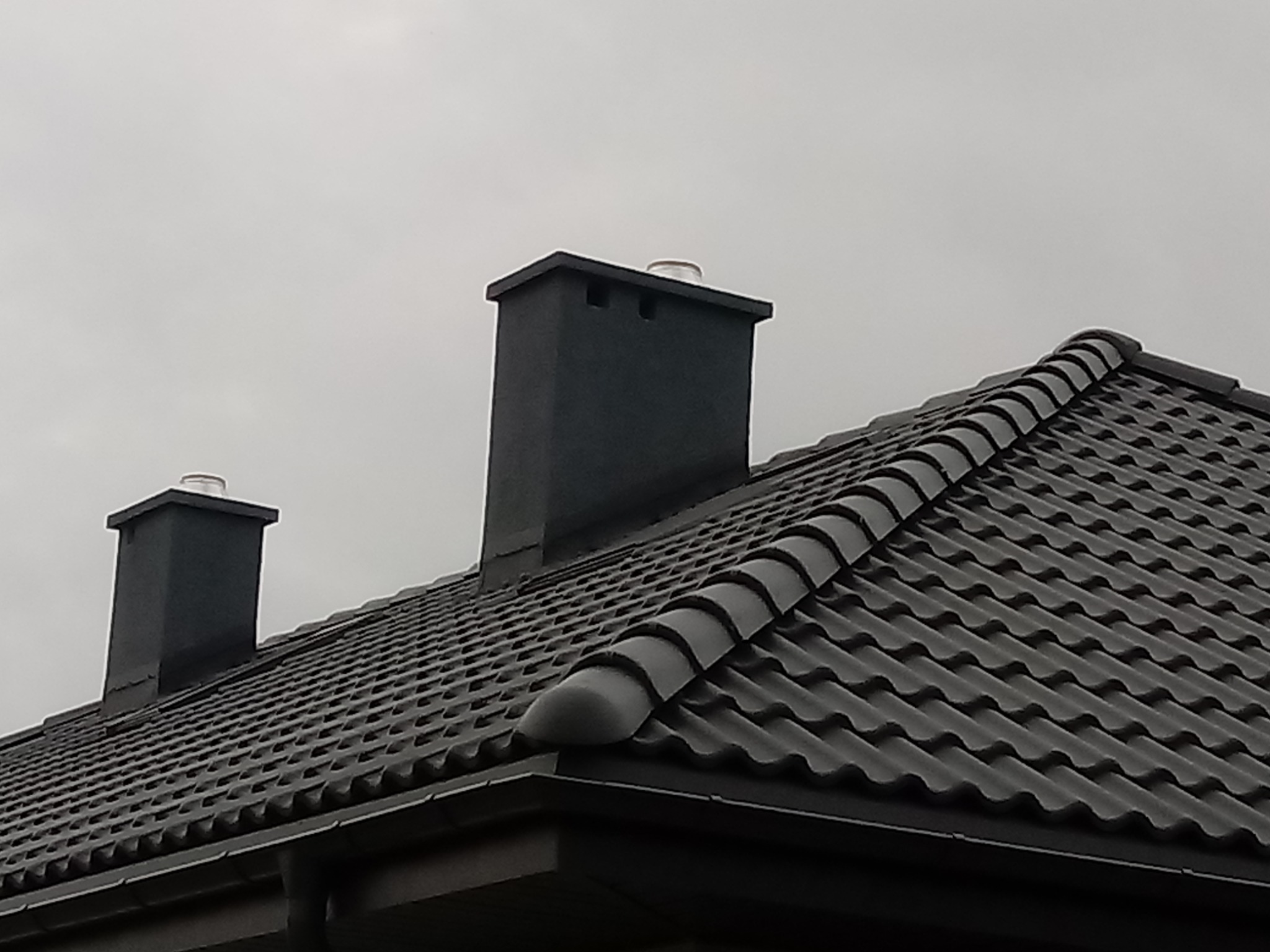 Grafitowy dach z dwoma kominami wentylacyjnymi i rynną, sfotografowany w pochmurny dzień.
