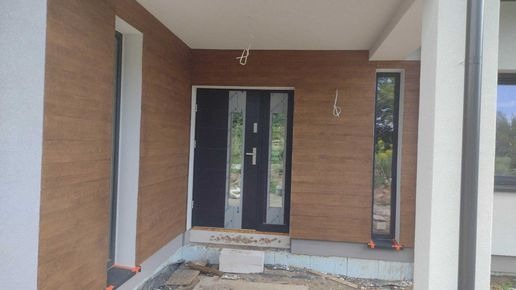 Nowe brązowe panele elewacyjne imitujące drewno na ścianie domu jednorodzinnego, czarne drzwi wejściowe z szybami, wąskie okno pionowe, widoczne kable wystające ze ściany, teren wokół w trakcie...