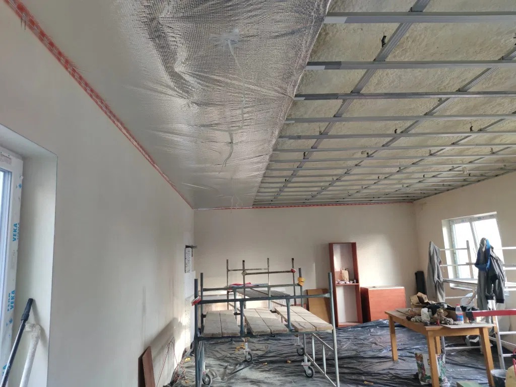 Wnętrze w trakcie prac: częściowo ocieplony sufit folią refleksyjną, metalowa konstrukcja podwieszana, ściany przygotowane do malowania, rusztowanie robocze.