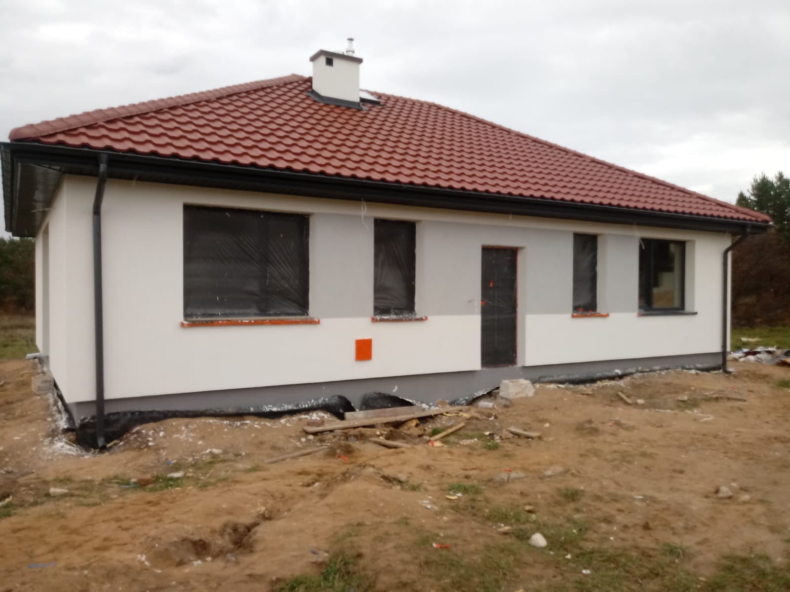 Nowo wybudowany dom jednorodzinny z czerwoną dachówką, z oknami zabezpieczonymi folią, częściowo otynkowany, z widocznymi elementami izolacji przy fundamentach i niezagospodarowanym terenem wokół.
