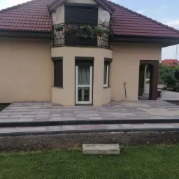 Nowo ułożony taras z szarej kostki brukowej przed domem z beżową elewacją i balkonem z kwiatami, widoczna łopata oparta o ścianę budynku.