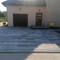 Ułożona, szara kostka brukowa przed domem z ciemnobrązową bramą garażową i drzwiami wejściowymi; widoczne obrzeża i fragment pracownika w lewym dolnym rogu.