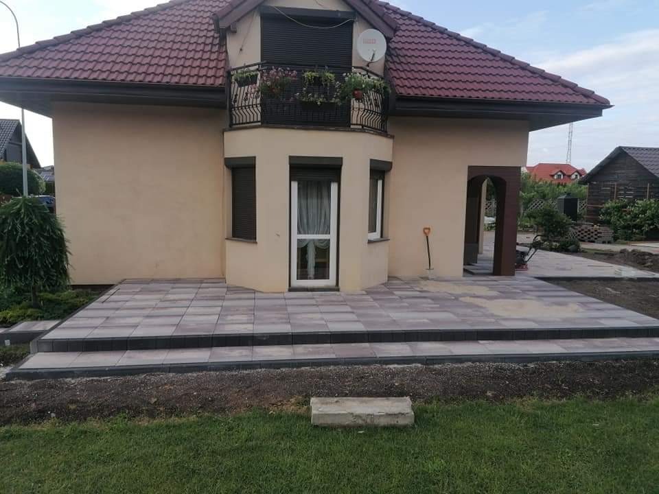 Nowo ułożony taras z szarej kostki brukowej przed domem z beżową elewacją i balkonem z kwiatami, widoczna łopata oparta o ścianę budynku.
