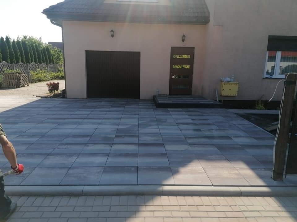 Ułożona, szara kostka brukowa przed domem z ciemnobrązową bramą garażową i drzwiami wejściowymi; widoczne obrzeża i fragment pracownika w lewym dolnym rogu.