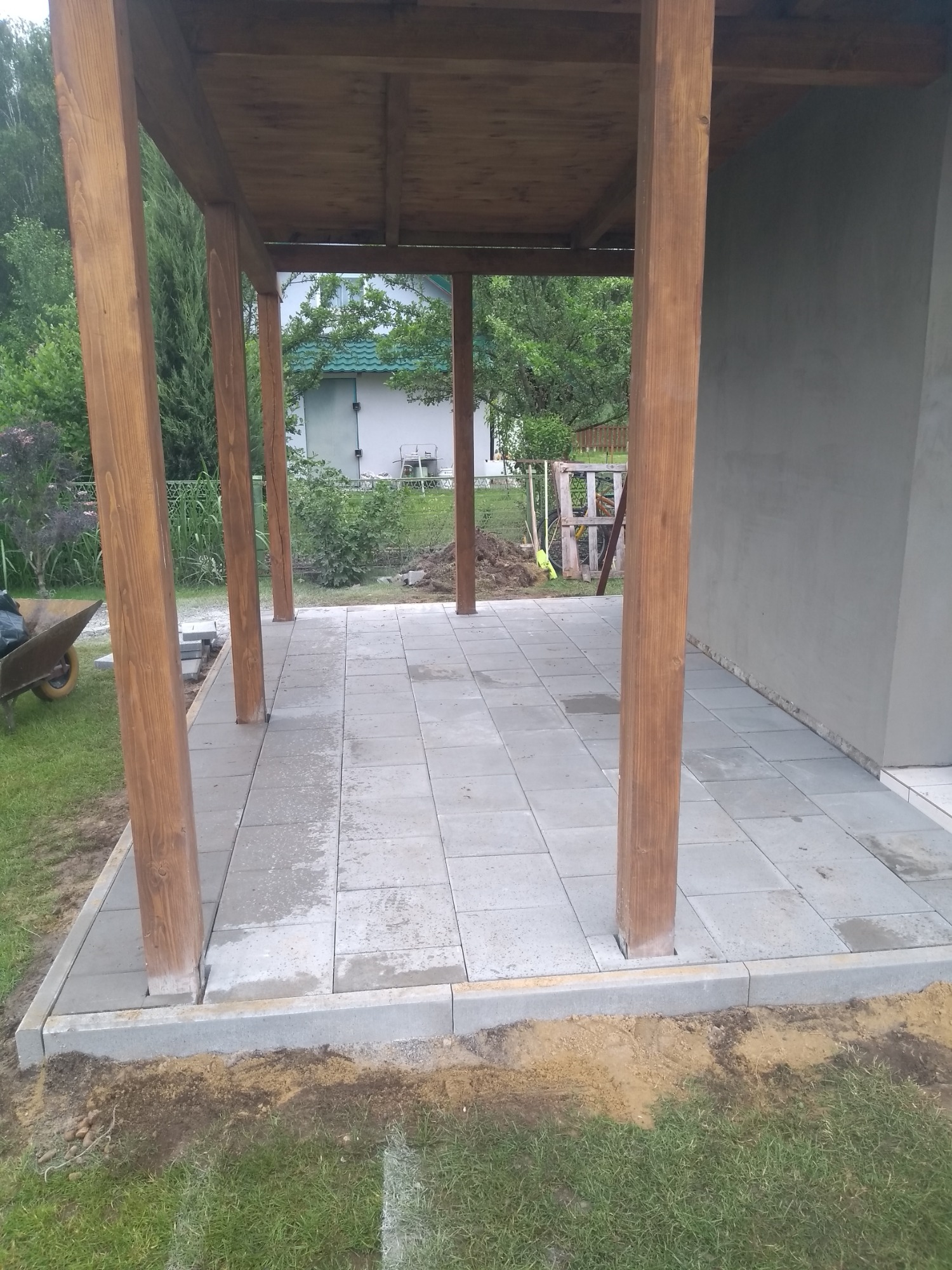 Konstrukcja z drewnianymi słupami i zadaszeniem, wyłożona szarymi płytami tarasowymi, widok na ogród z wózkiem i dom w tle.