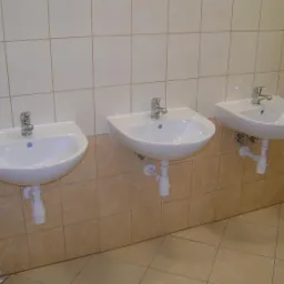 Trzy białe umywalki zamontowane na ścianie z beżowych płytek, widoczne instalacje wodno-kanalizacyjne.