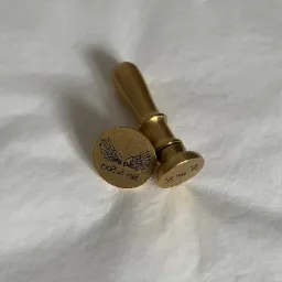 Złoty stempel do laku z drewnianą rączką i okrągłą pieczęcią z motywem skrzydeł i napisem 'Sky & Soul', leżący na białym tle.