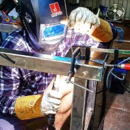 Spawanie metalowego elementu konstrukcyjnego w warsztacie, widoczna maska spawalnicza, rękawice ochronne i zaciski stolarskie mocujące spawany profil.