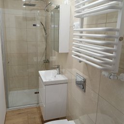 Alex-Tech - Nowoczesna łazienka z beżowymi płytkami, kabiną prysznicową typu walk-in, białą szafką z umywalką, wiszącym grzejnikiem drabinkowym i podłogą z paneli imitujących drewno.