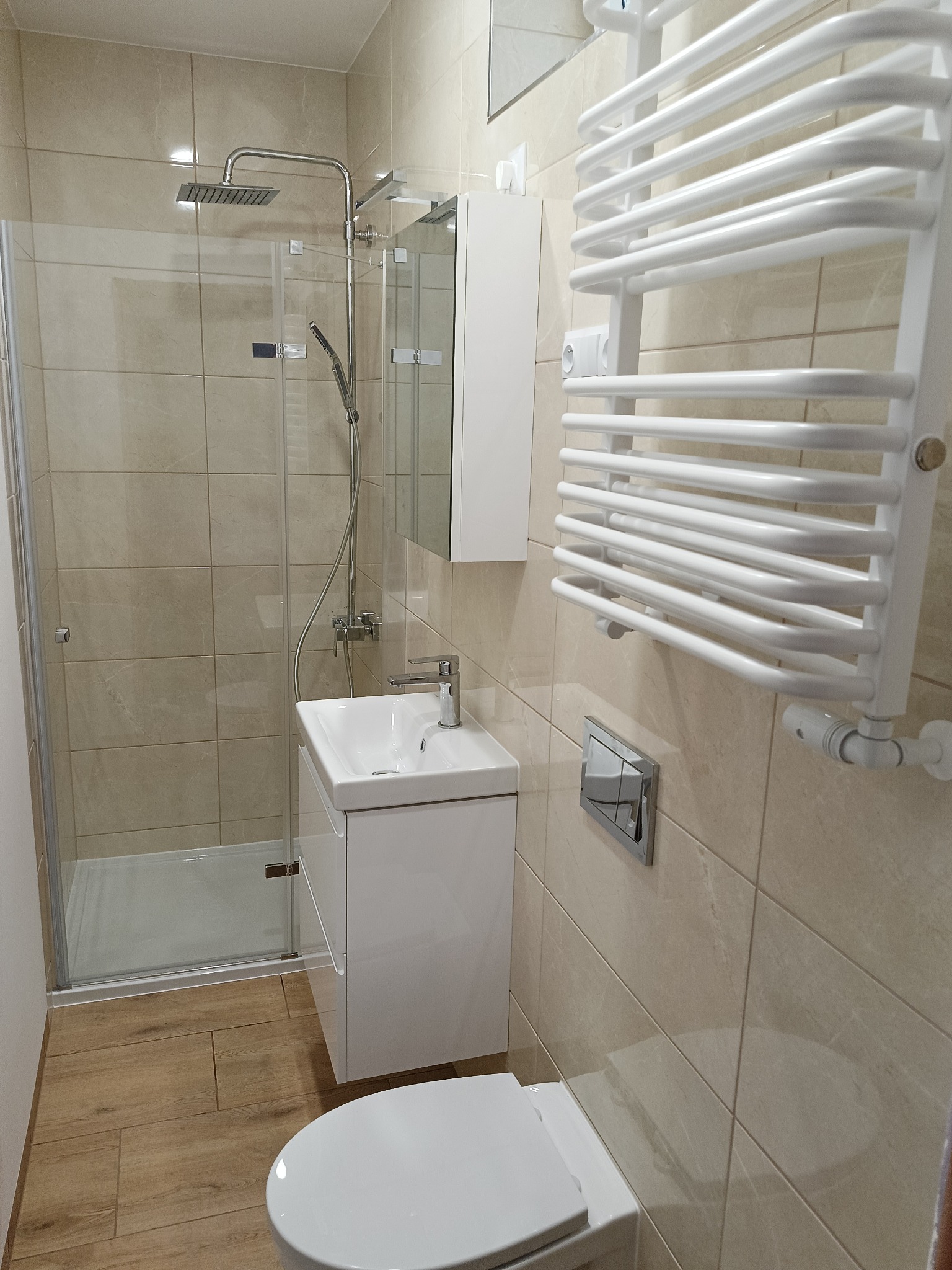 Nowoczesna łazienka z beżowymi płytkami, kabiną prysznicową typu walk-in, białą szafką z umywalką, wiszącym grzejnikiem drabinkowym i podłogą z paneli imitujących drewno.