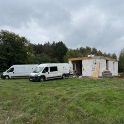 Przedsiebiorstwo Wielobranżowe Rafal Zadworny - Budowa parterowego domu z betonu komórkowego, widoczne dwa busy firmy na tle zieleni i pochmurnego nieba. Etap murowania ścian.