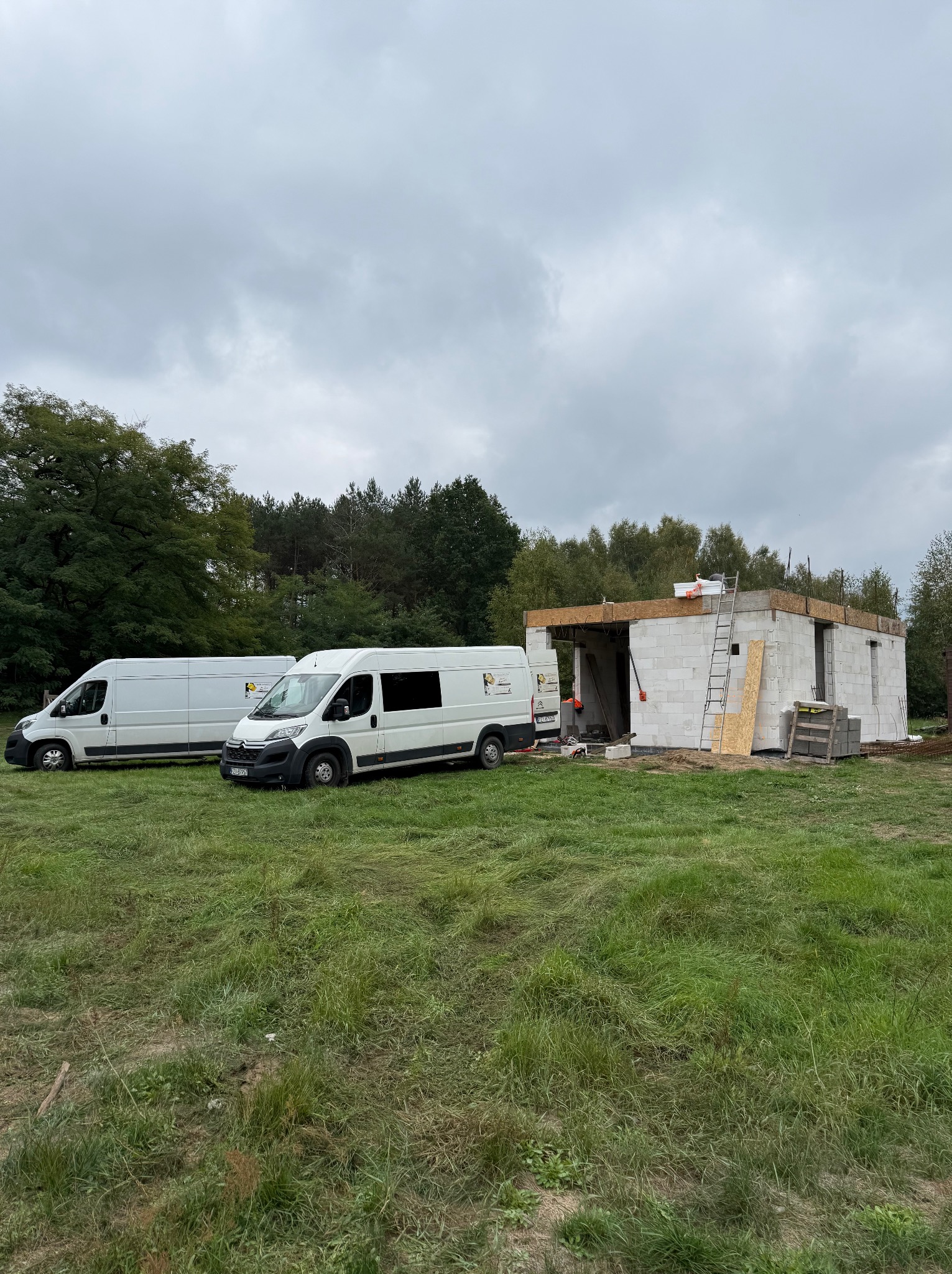 Budowa parterowego domu z betonu komórkowego, widoczne dwa busy firmy na tle zieleni i pochmurnego nieba. Etap murowania ścian.