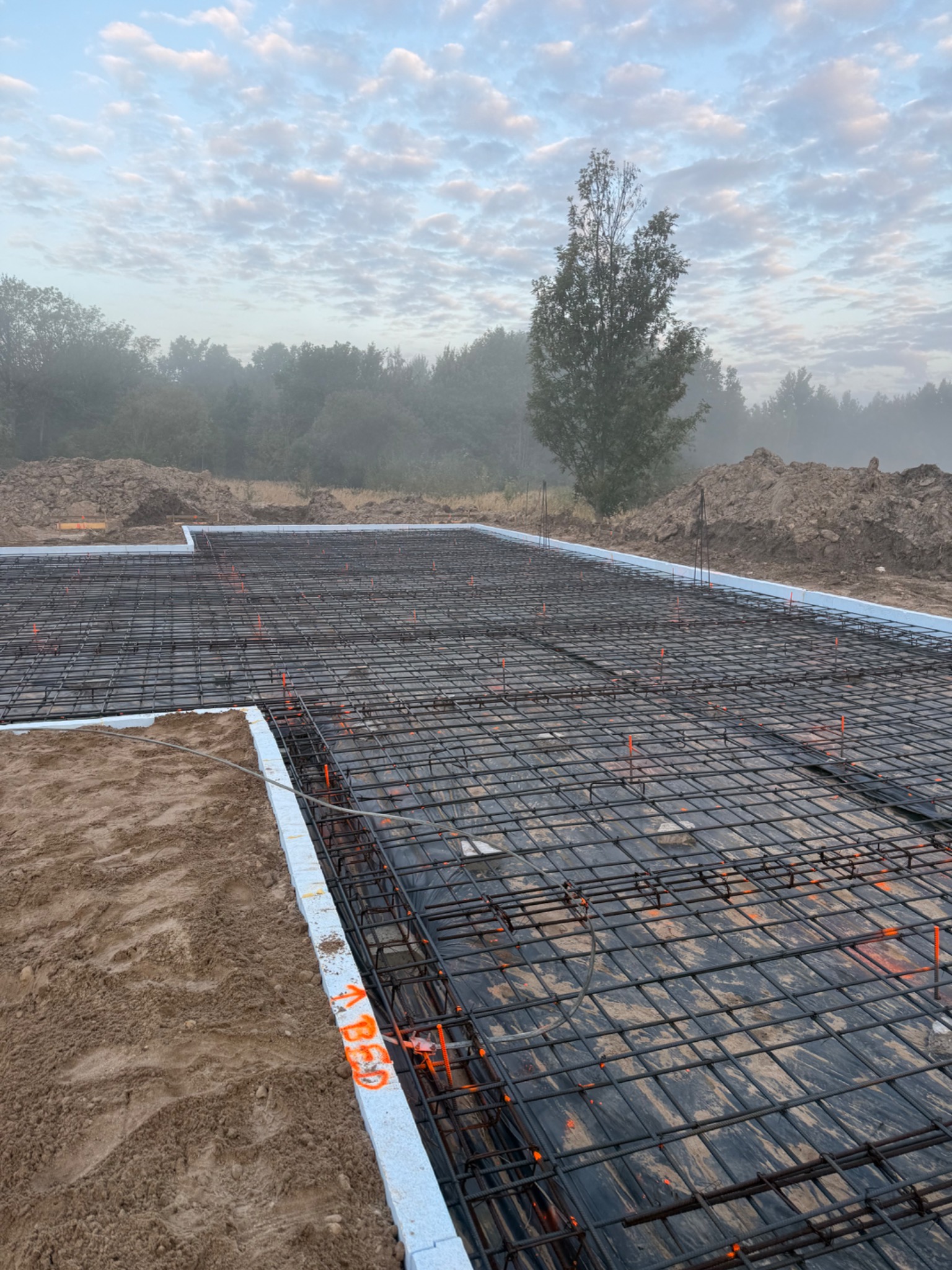 Zbrojenie fundamentów pod dom w Świdnicy, widok z góry. Stalowe pręty tworzą siatkę na czarnej folii, obok białe bloczki z pomarańczowymi oznaczeniami.