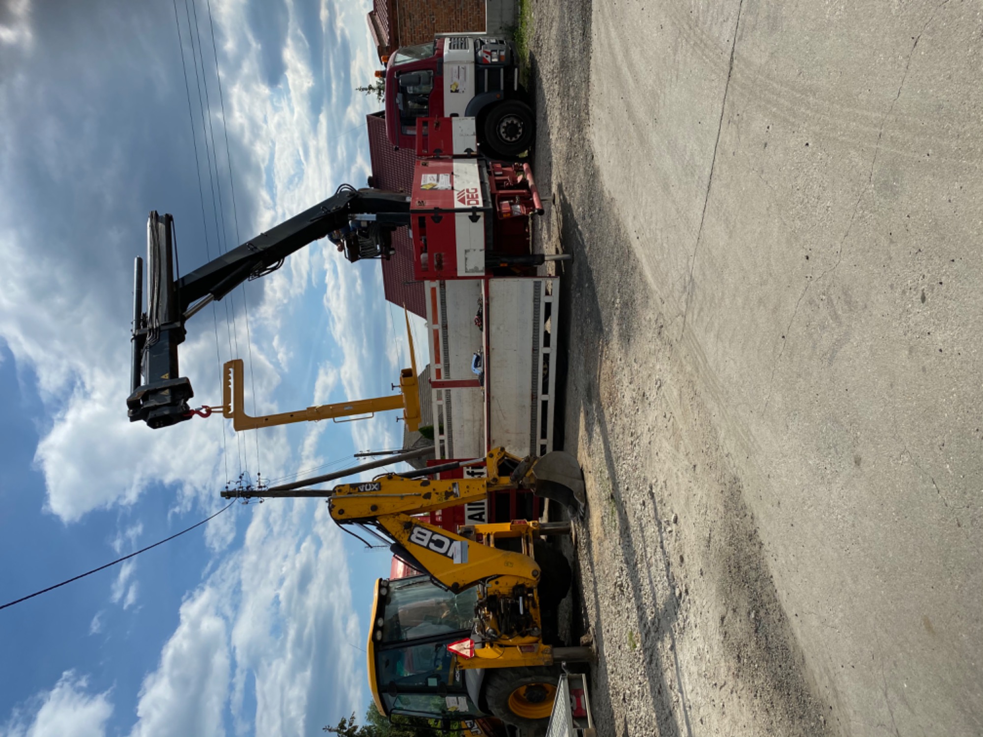 Żółta koparko-ładowarka JCB i ciężarówka z dźwigiem na poboczu drogi, widoczne słupy energetyczne i błękitne niebo z chmurami.