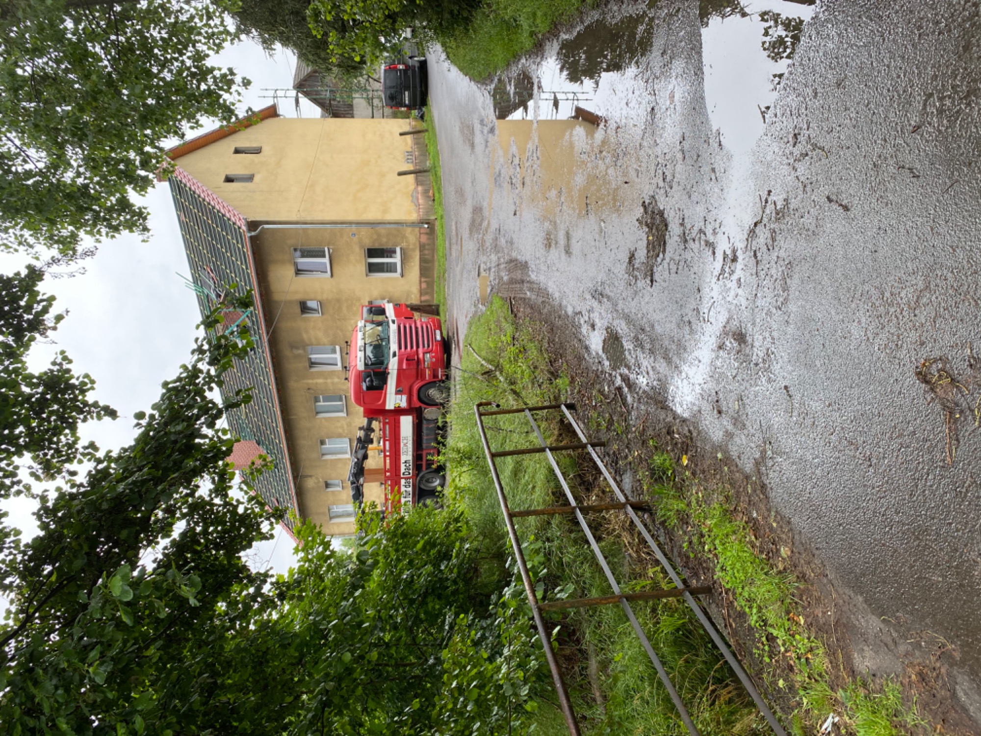 Żółty budynek z częściowo zamontowaną konstrukcją dachu, widoczny z mokrej drogi z kałużami, obok zaparkowany czerwony samochód ciężarowy z dźwigiem, w tle czarny samochód osobowy.