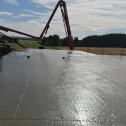 Świeżo wylany, mokry beton na płycie fundamentowej domu jednorodzinnego, widoczna pompa do betonu, wystające pręty zbrojeniowe i szalunki, w tle zabudowa jednorodzinna i las.