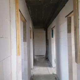 Wnętrze surowego stanu budynku z białych bloczków, widoczne otwory na drzwi z drewnianymi ościeżnicami, betonowa posadzka i sufit, perspektywa korytarza w głąb budynku.