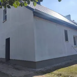 Dom w Chociwlu po nałożeniu pierwszej warstwy elewacji w kolorze jasnoszarym, widoczny fragment dachu z brązową dachówką i niezabudowany otwór drzwiowy.