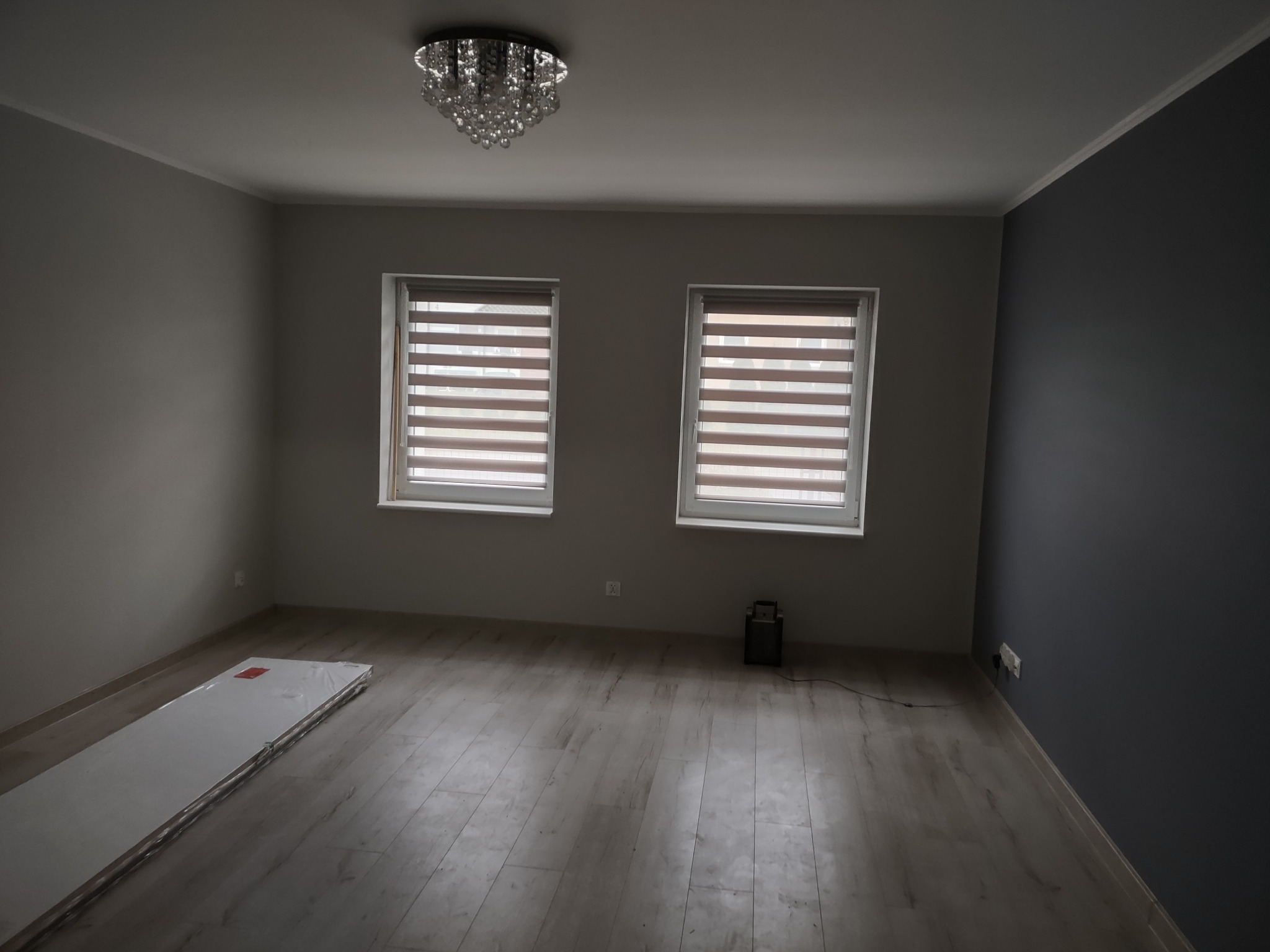 Pusty pokój po remoncie z nowymi panelami podłogowymi, roletami w oknach i eleganckim żyrandolem, przygotowany do dalszego wykończenia.