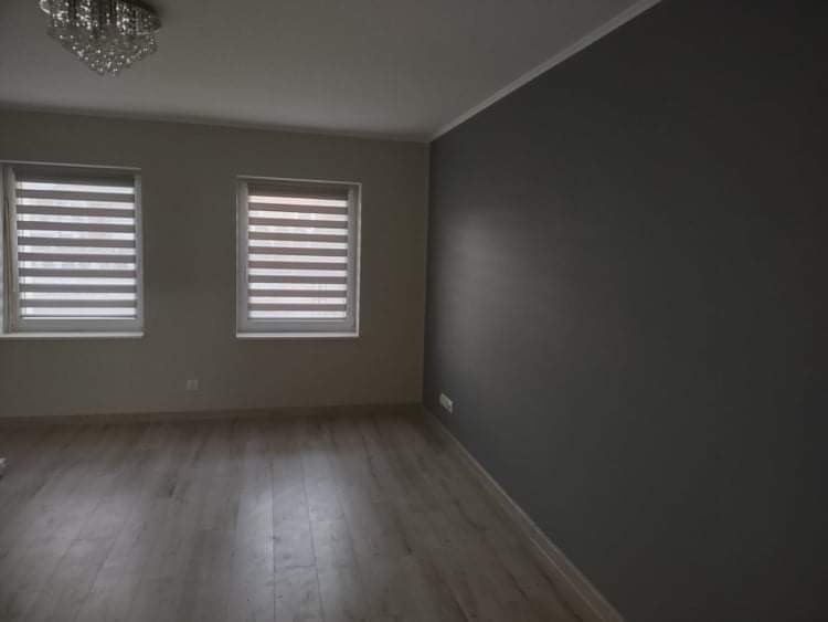 Pomalowane na szaro wnętrze pokoju z drewnianą podłogą, dwoma oknami z żaluzjami poziomymi i ozdobnym żyrandolem na suficie.