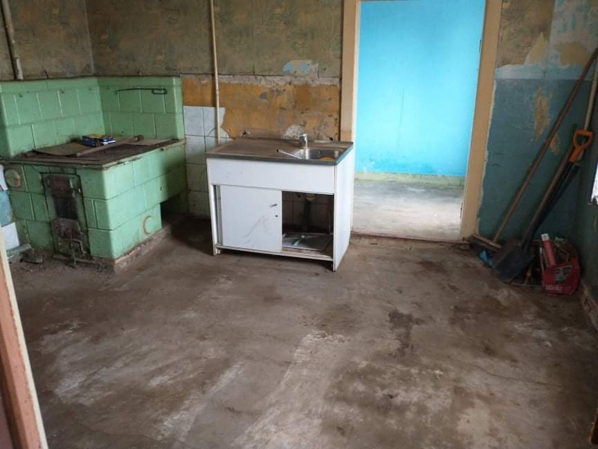 Wnętrze z piecem kaflowym i umywalką przed remontem, widoczne ślady wilgoci na ścianach i podłodze, narzędzia opierają się o ścianę w tle.