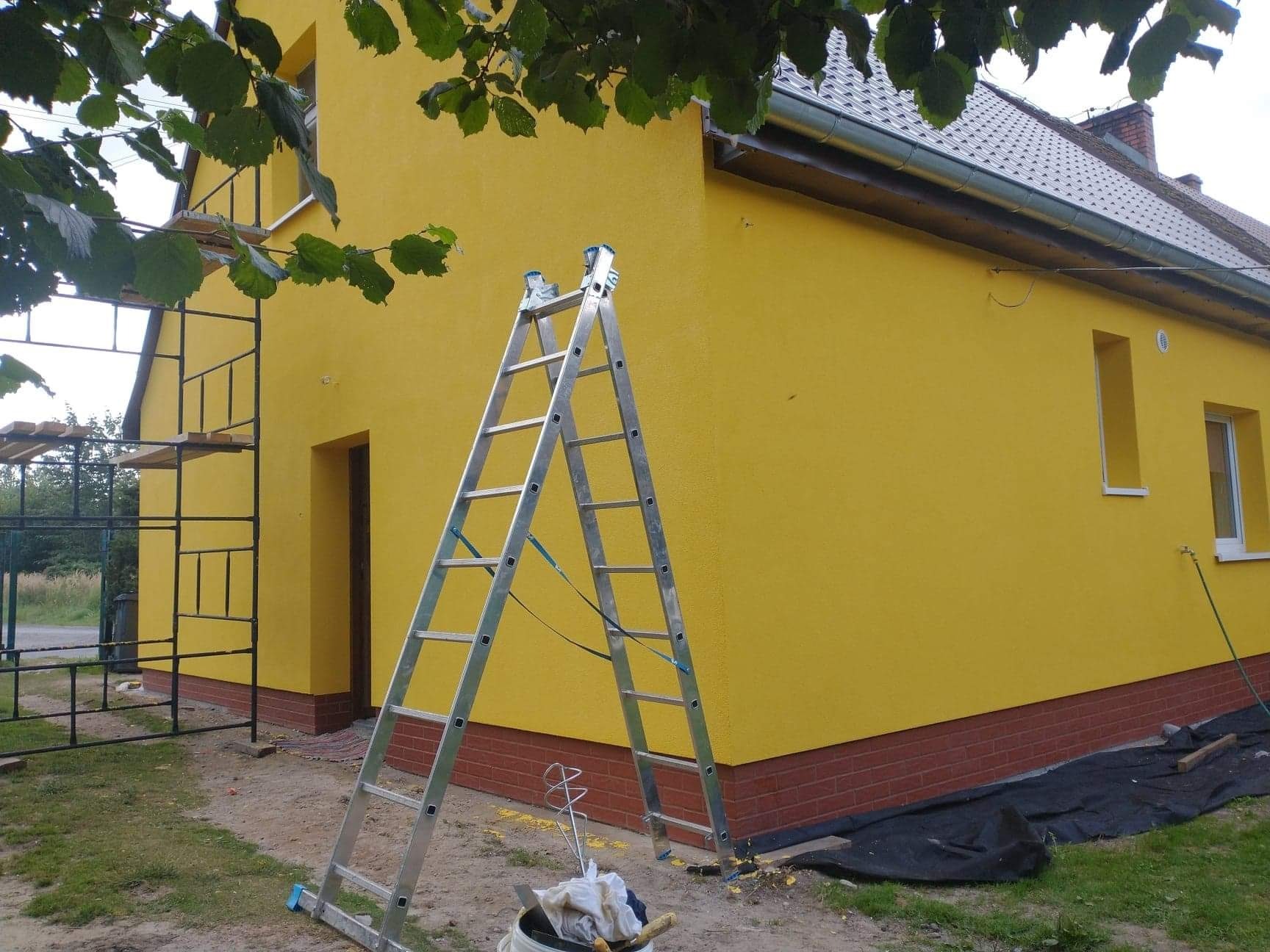 Żółta elewacja domu jednorodzinnego w trakcie prac, widoczne rusztowanie, drabina i narzędzia malarskie.