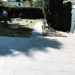 Widok na ogr&oacute;d z nowo ułożoną, prostokątną, szarą nawierzchnią z kostki. W tle trawnik z okrągłym, betonowym elementem, pracownicy z taczką i świeżo przygotowane rabaty.