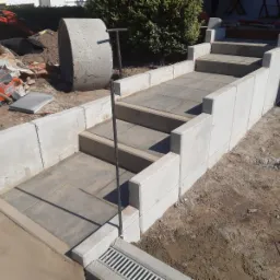 Betonowe schody z kostki brukowej w trakcie budowy, widoczne materiały budowlane, piła do cięcia kostki oraz rura betonowa w tle.