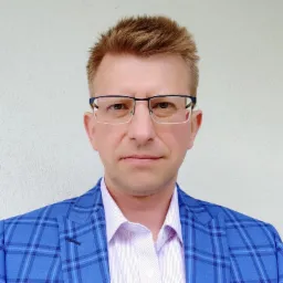 Portret mężczyzny w okularach i niebieskiej marynarce w kratę na tle jasnej ściany.