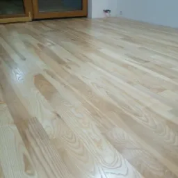 Świeżo położona, jasna podłoga z desek parkietowych, widoczne naturalne usłojenie drewna, w tle drewniane drzwi i biała ściana z gniazdkiem elektrycznym.