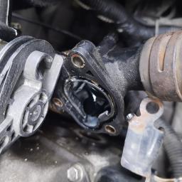 Daniel Bzowski - Zdjęcie zbliżeniowe demontażu termostatu samochodowego z widocznymi śladami korozji na elementach mocujących.