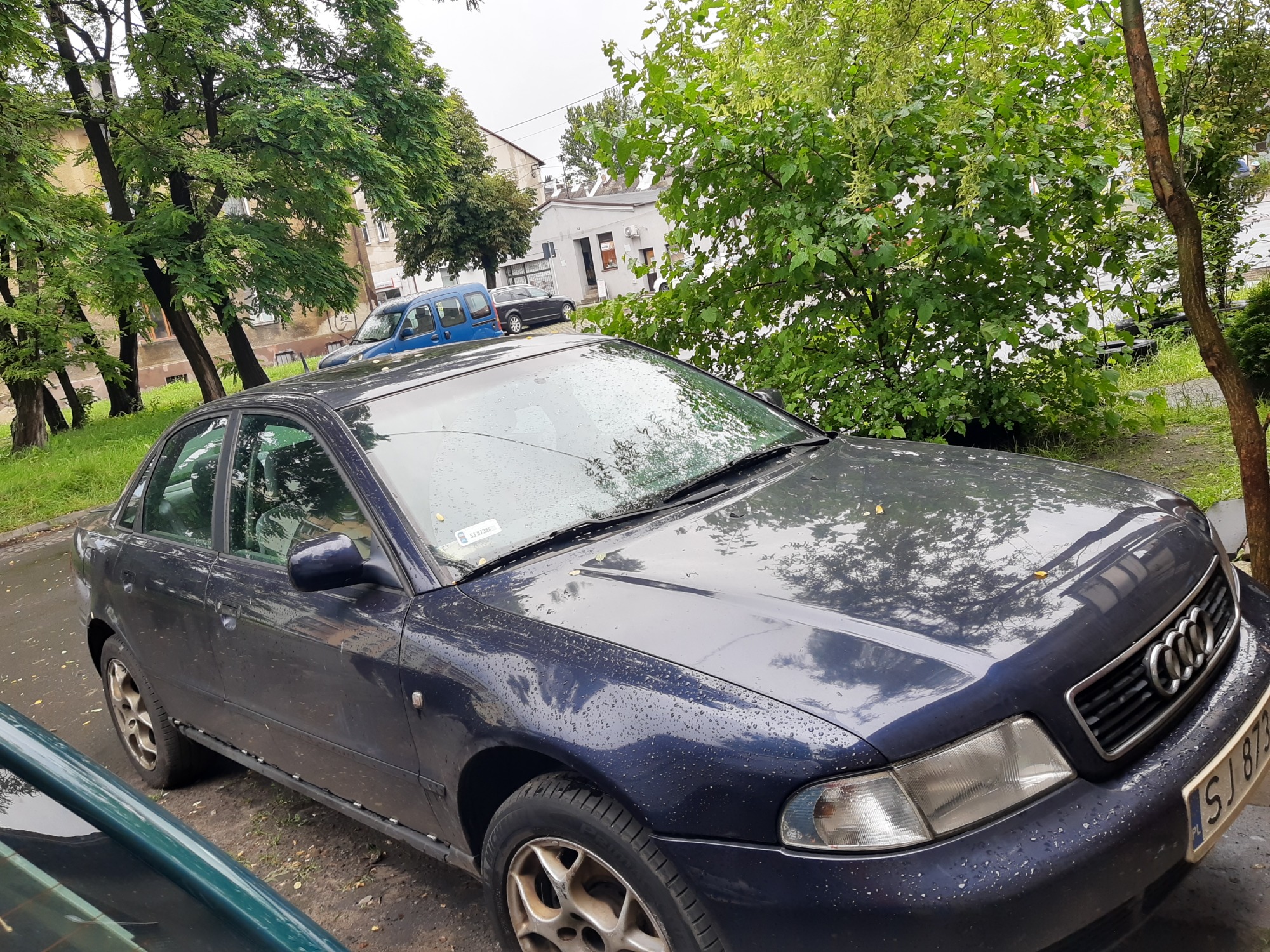 Granatowe Audi A4 na parkingu, widoczne ślady deszczu na karoserii, w tle budynek i drzewa.