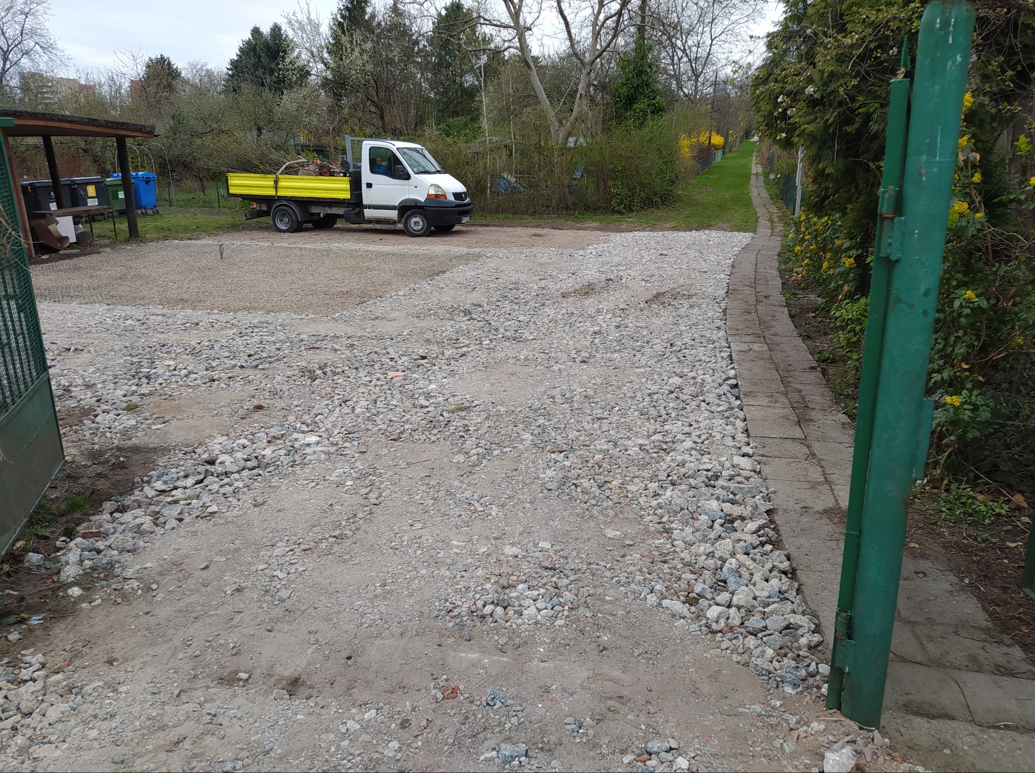Świeżo wysypany żwir na podwórku z widocznym samochodem dostawczym z żółtą skrzynią ładunkową, zielona brama wjazdowa po prawej stronie i częściowo utwardzona droga z kostki brukowej prowadząca...