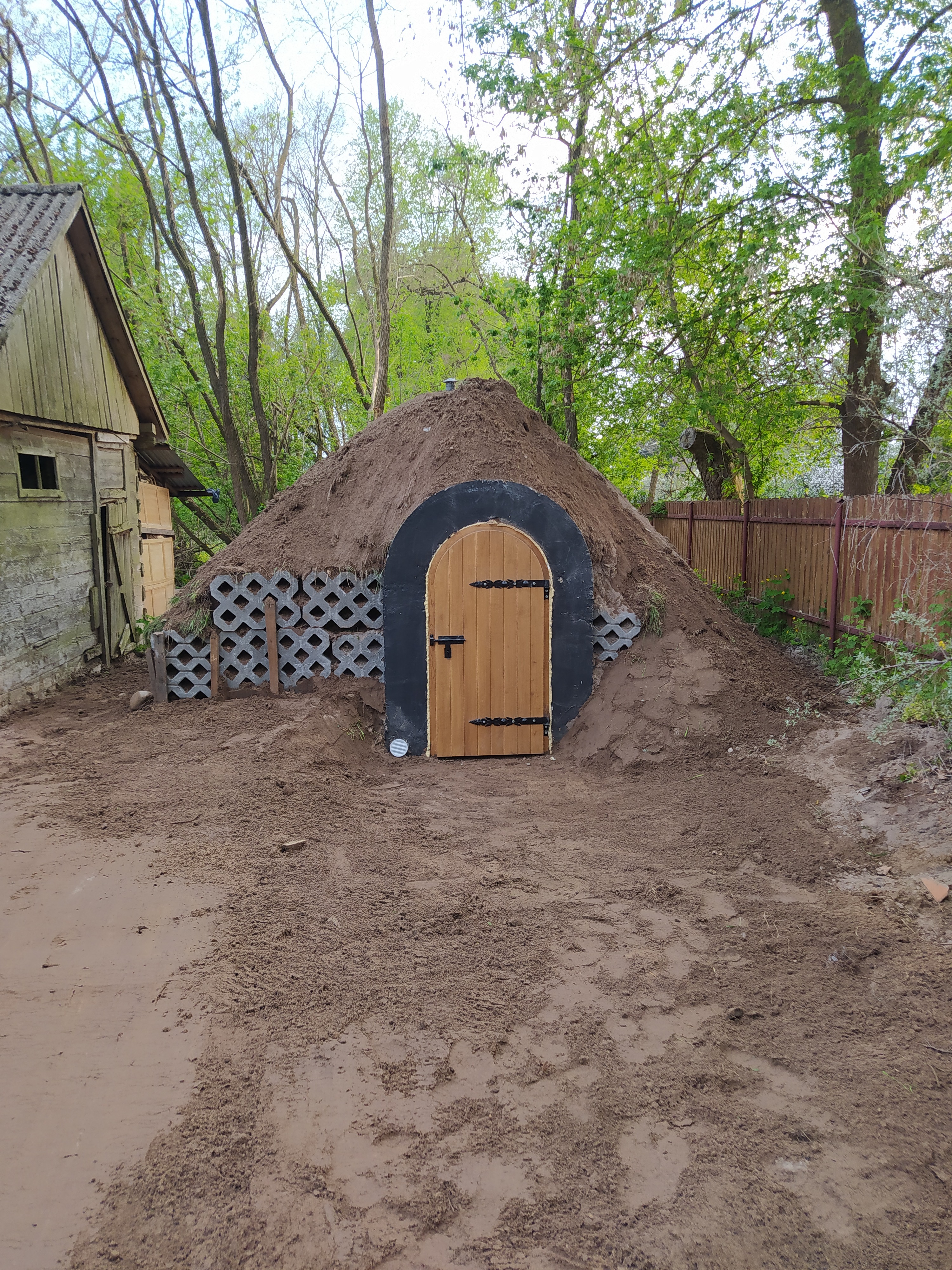 Ziemianka z drewnianymi drzwiami z czarnymi okuciami, wzmocniona betonowymi ażurami, otoczona ziemią i drzewami.